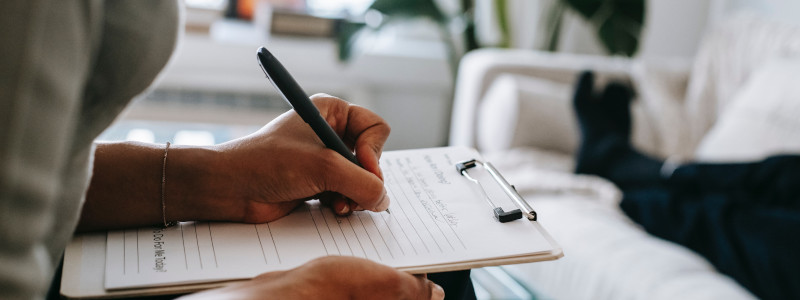 An image showing a hypnotherapist working with a male patient. You can only see the hypnotherapists hands holding a pen writing on notes on a  clipboard (on their lap), and you can only see the patient's black trousers, and black socked feet, lying on a white couch.